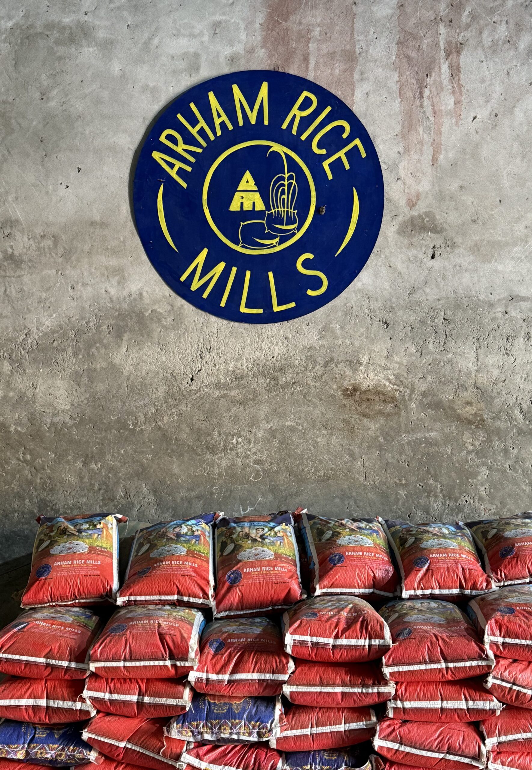 Stacked premium basmati rice bags under Arham Rice Mills logo on warehouse wall